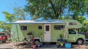 Een groene camper met een afgedekt voorportaal, een klein terrasje met zitplaatsen en diverse potplanten.