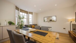 Un área de comedor con una mesa de madera, sillas grises, un sofá con cojines, un ventanal grande y un espejo en la pared.