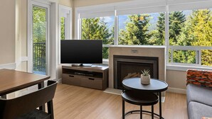 A living room with a fireplace, a television, and a dining area with a table and chairs. - Christmas in Whistler!   1 bedroom + den condo. Walk to the slopes!  (Whistler)
