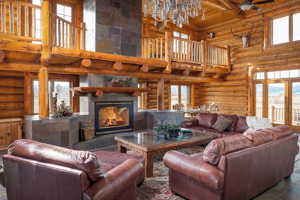 A log cabin interior with a fireplace, leather sofas, a glass coffee table, and a balcony with a railing.