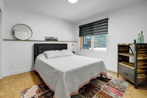 A bedroom with a bed, a window with blinds, a mirror, a small shelf with bottles, and a patterned rug.