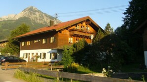 Un edificio de madera con varias ventanas y balcones, rodeado de vegetación y una montaña al fondo.