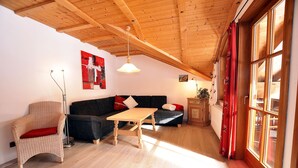 Una habitación acogedora con techo de madera, un sofá oscuro, una silla de mimbre, una mesita y una ventana con cortinas.