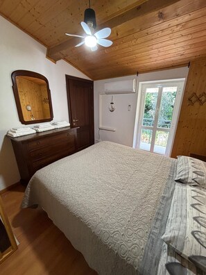 A bedroom with a wooden ceiling, a ceiling fan, a bed with a patterned bedspread, a dresser, a mirror, and a window with a view of greenery. - The Spiral House (Liguria)