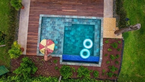 Piscina retangular com um deck de madeira ao redor, uma pequena mesa redonda com um ventilador, e uma área gramada com plantas.