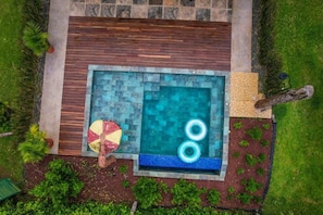 Una piscina rectangular con un deck de madera que la rodea, una pequeña mesa redonda con un ventilador y una zona ajardinada con plantas.