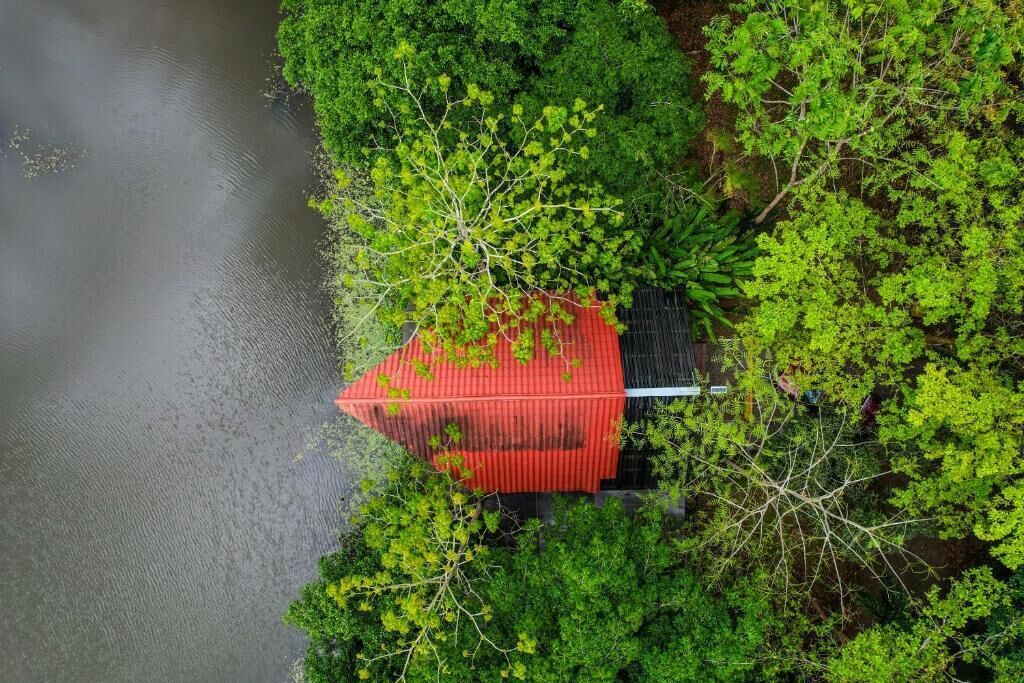 A red-roofed building surrounded by dense green foliage, adjacent to a body of water.