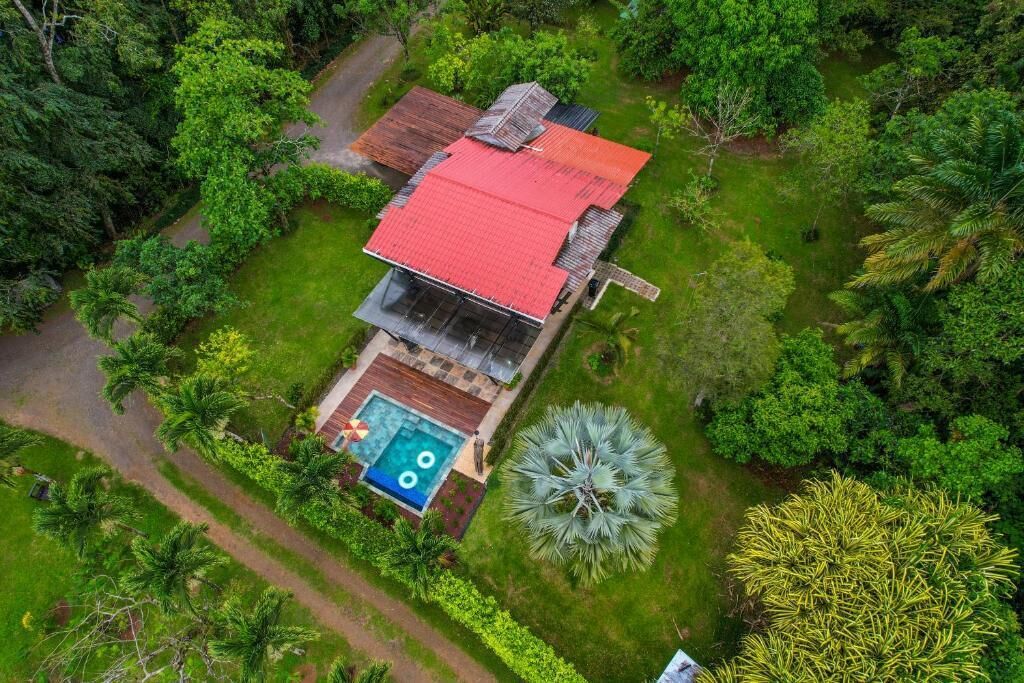 Una casa de un piso con techo rojo, rodeada de exuberante vegetación y una piscina.