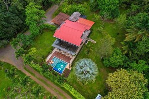 Una casa de un piso con techo rojo, rodeada de exuberante vegetación y una piscina.