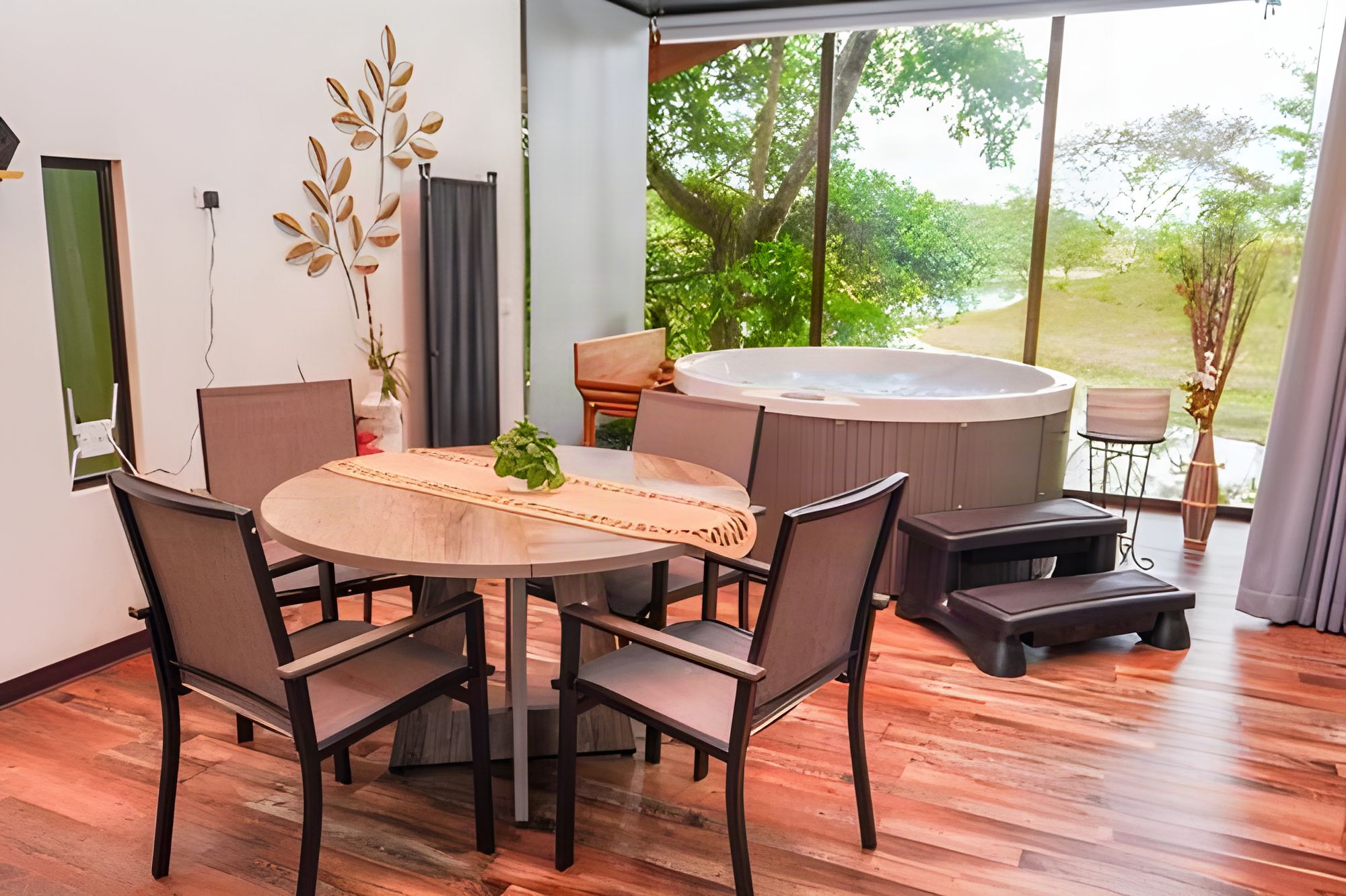 A modern indoor space with a round wooden table, dark chairs, a hot tub, and a view of a lush garden.