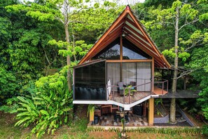 Una casa en el árbol con cerramiento de cristal, terraza de madera y mobiliario exterior.