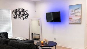 A modern living room with a sectional sofa, a flat-screen TV mounted on the wall, a decorative wall piece, and a small round table with a candle and figurine.