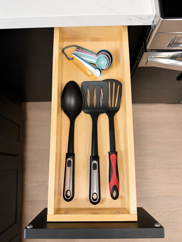 A kitchen drawer with a wooden interior, containing a black ladle, a black spatula with red handle, and a set of measuring cups.