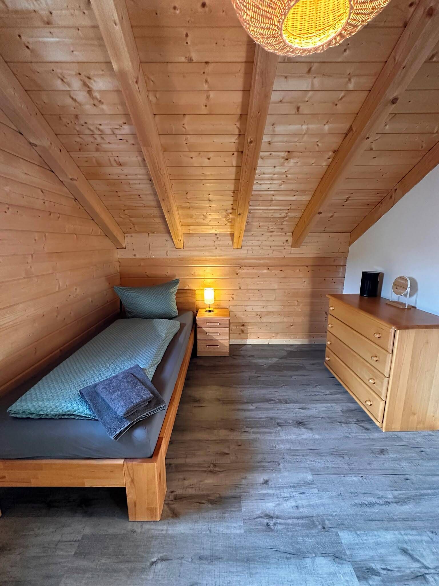 Et soveværelse i træ på loftet med seng, natbord, kommode og hængende lampe.