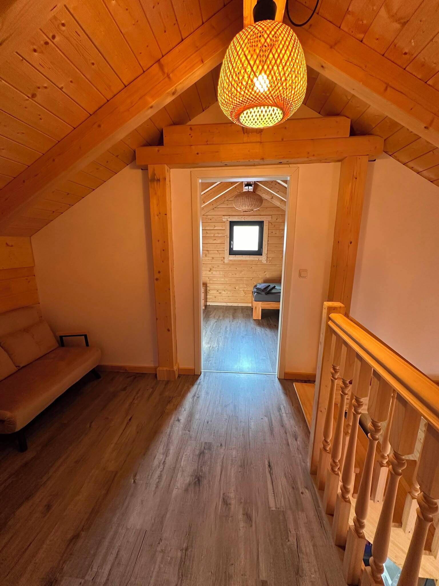 A wooden interior with a sloped ceiling, a sofa, a hanging light fixture, and a doorway leading to another room.