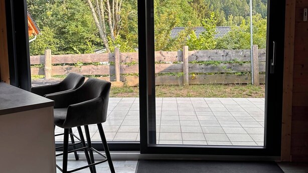 Et moderne interiør med træloft, en skydedør til en udendørs terrasse og et lille siddeområde med stole.