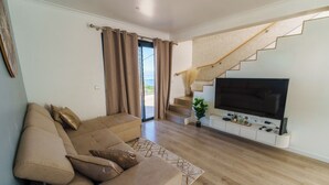 A modern living room with a sofa, a flat-screen TV, a staircase, and a view to the outside.