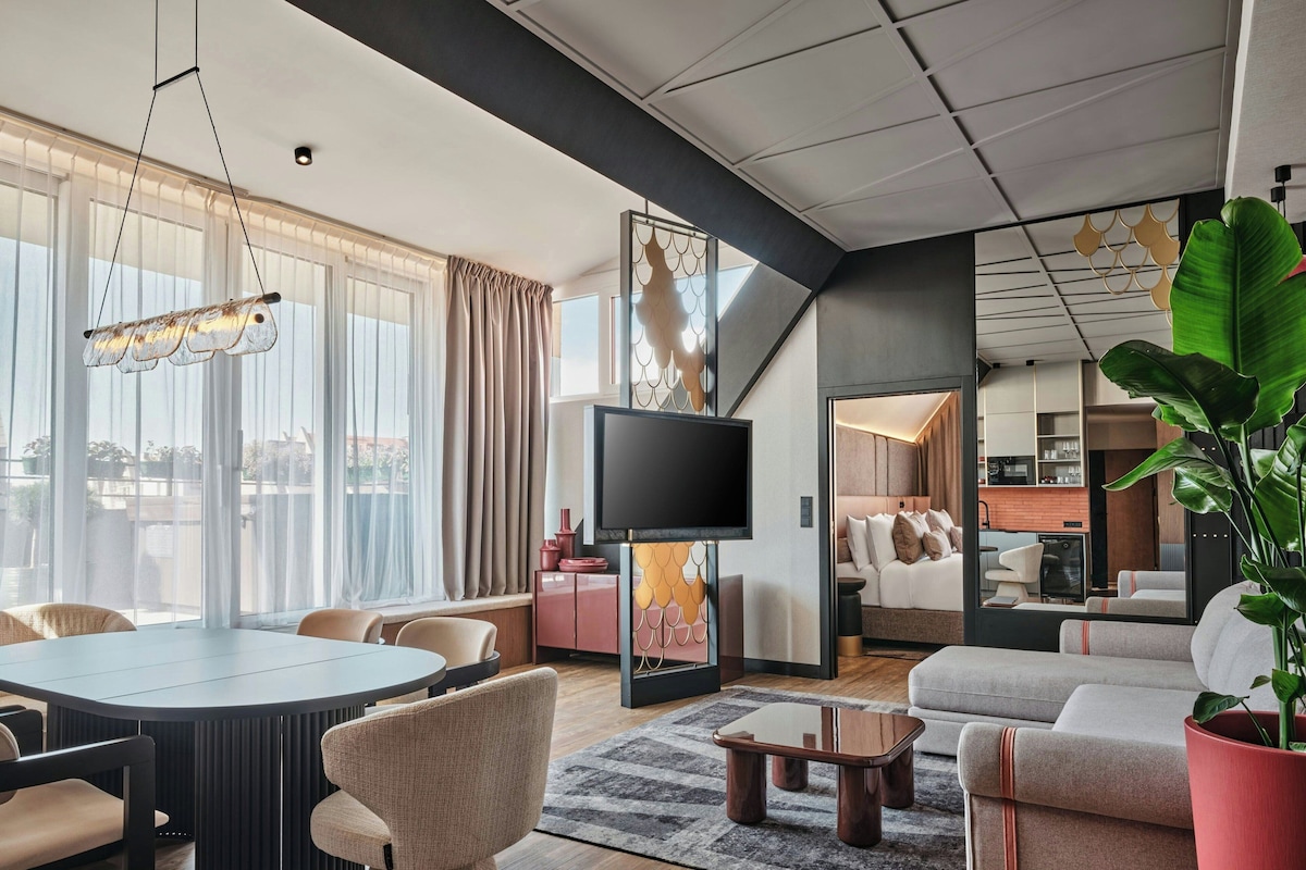 Una habitación de hotel moderna con comedor, televisor de pantalla plana, sofá y una planta grande.