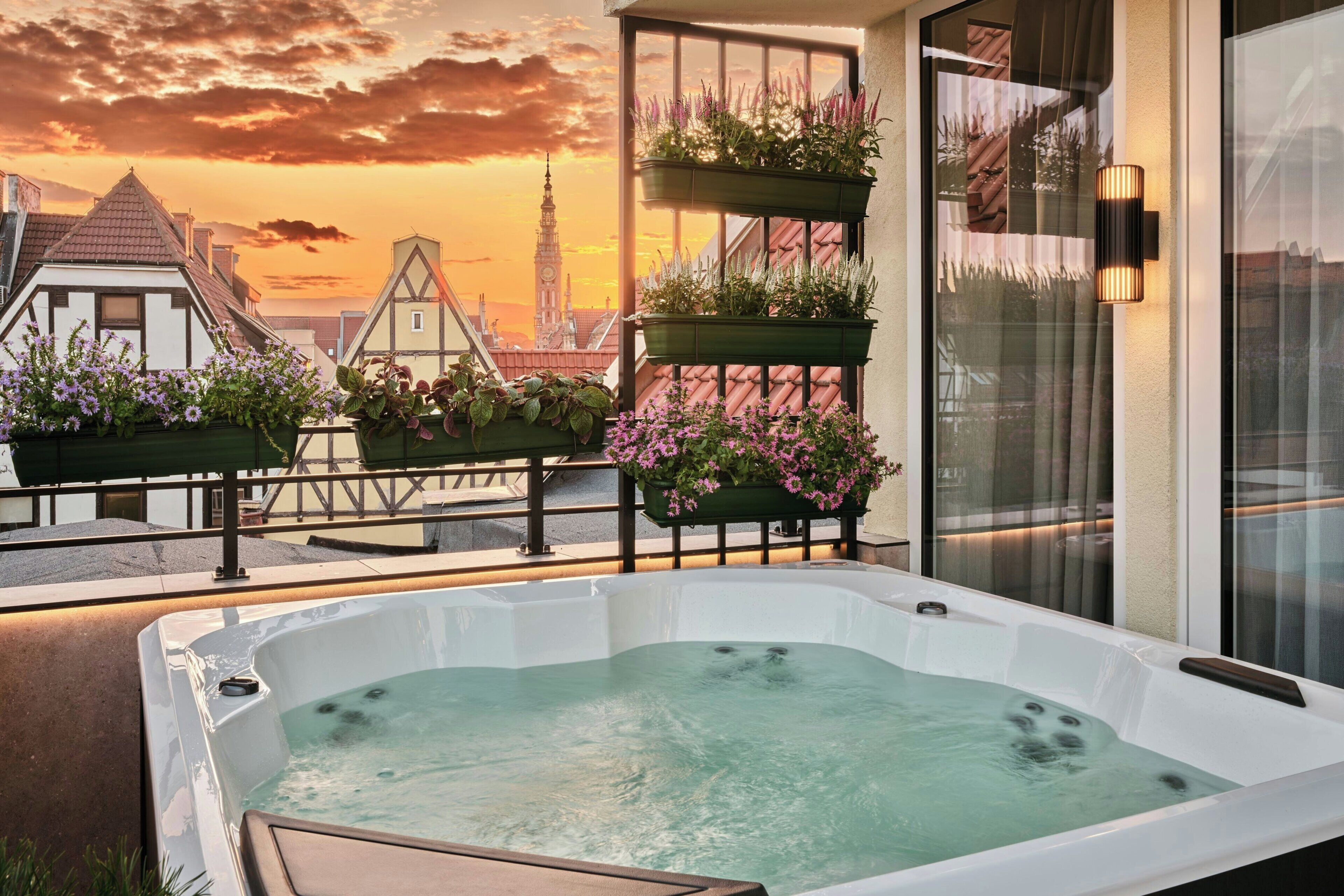 A hot tub on a balcony with a cityscape view, potted plants, and a modern railing.