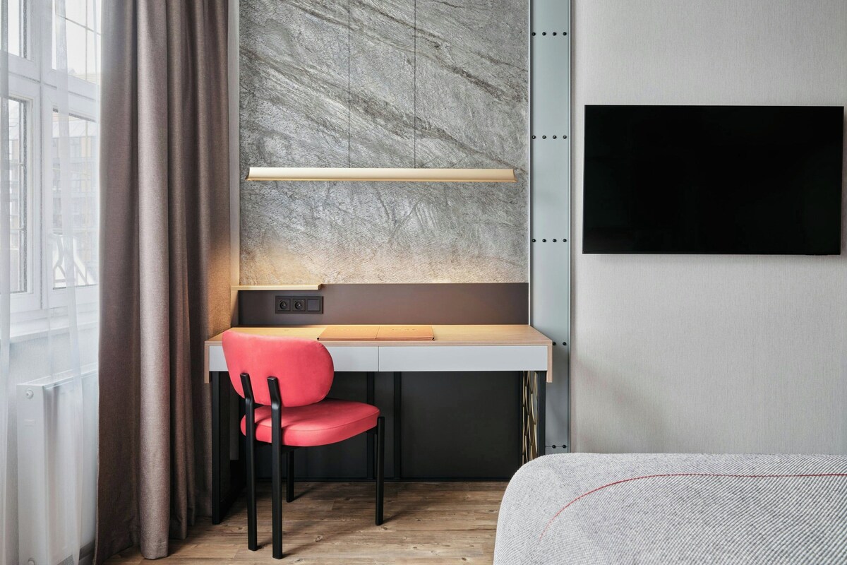 A modern hotel room with a desk, a red chair, a flat-screen TV, and a large window with curtains.