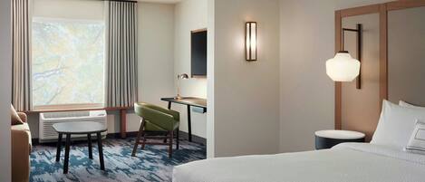 Ein Hotelzimmer mit Bett, Schreibtisch, Stuhl, Lampe und Blick auf Bäume durch das Fenster.
