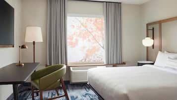 A hotel room with a bed, a desk with a lamp, a chair, a window with curtains, and a nightstand with a lamp.