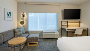 A hotel room with a sofa, ottoman, round table, desk with chair, TV, air conditioning unit, and artwork on the wall. - Towneplace Suites By Marriott Little Rock Otter Creek (Little Rock)