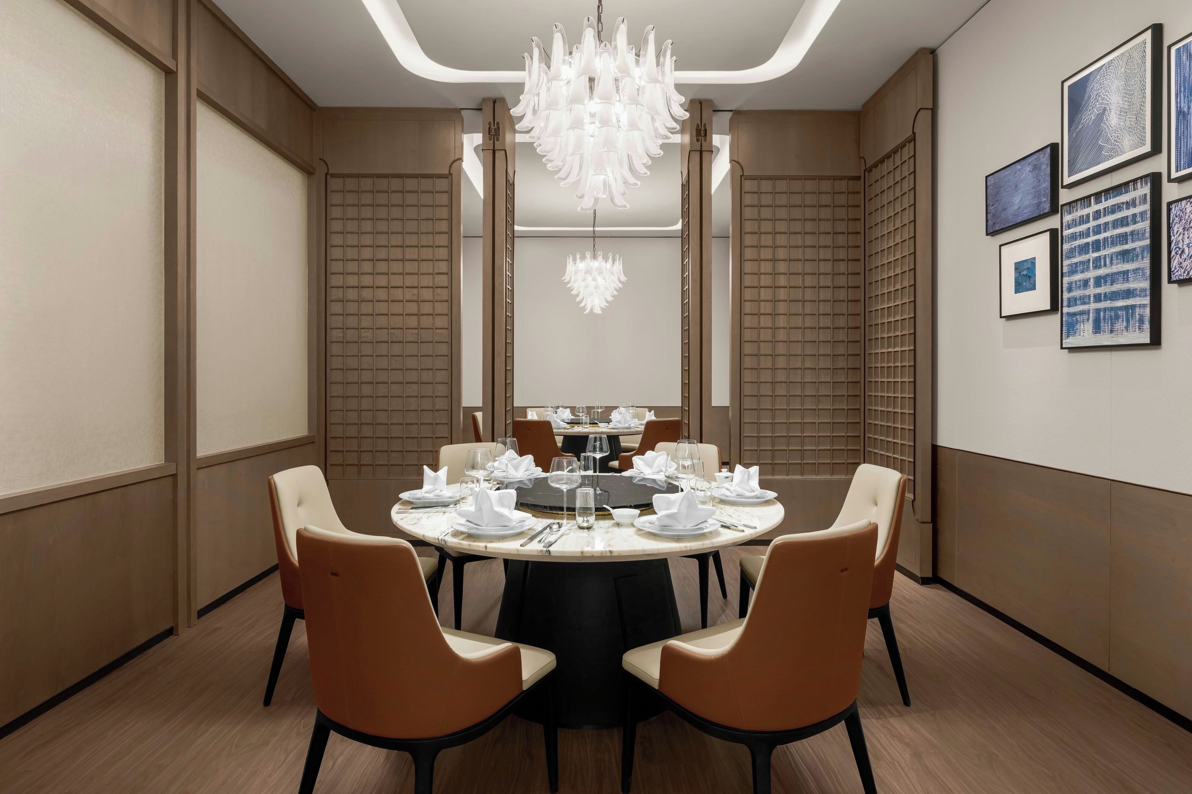 A dining area with a round table set for six, surrounded by brown chairs, with a chandelier above and artwork on the walls.