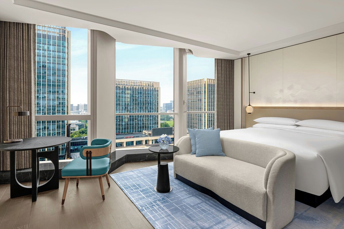 A modern hotel room with a large bed, a desk, a chair, and a view of the cityscape.