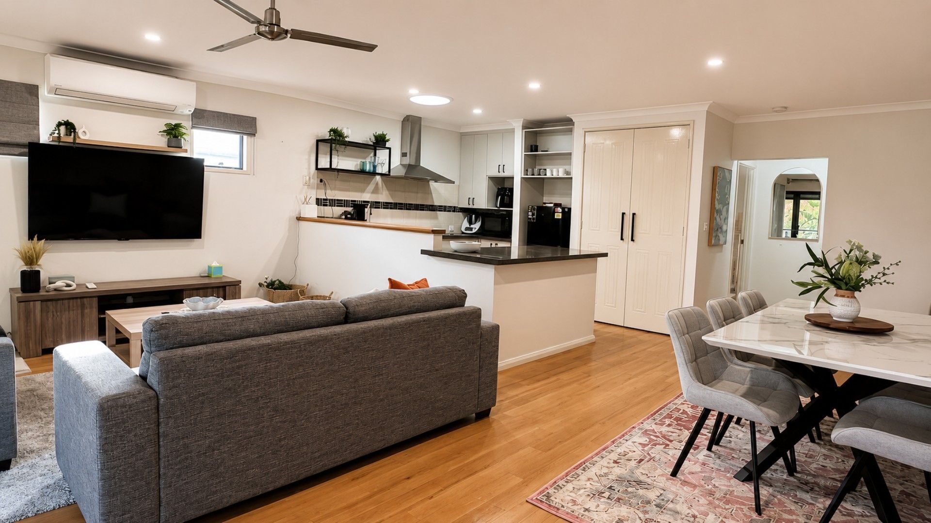 Una cocina moderna con televisor de pantalla plana, sofá gris, mesa de comedor con sillas y ventilador de techo.