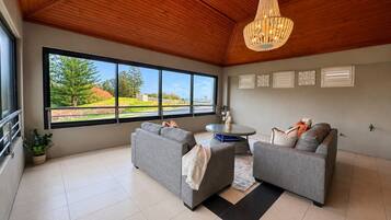 A spacious living room with a large window, grey sofas, a round table, and a chandelier.