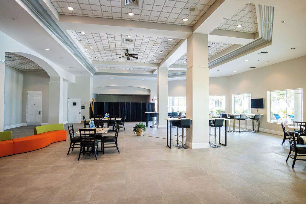 Um lobby espaçoso com teto alto, área de estar com almofadas laranja e verde, e um espaço de bar com banquetas e televisão.