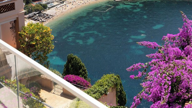 Blick aufs Meer von einem Balkon, darunter ein Strand, klares blaues Wasser und ein Gebäudekomplex im Hintergrund.