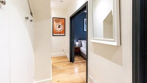 Un pasillo con piso de madera, una cama con ropa blanca, una mesita de noche con una lámpara y un cartel en la pared.