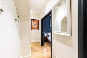 A hallway with wooden flooring, a bed with white bedding, a nightstand with a lamp, and a poster on the wall. - Modern Covent Garden Apartment (London)
