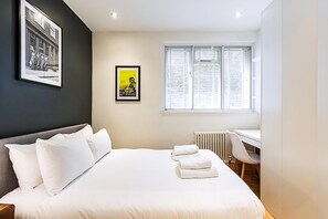 A bedroom with a bed, a desk, a chair, a radiator, and two framed pictures on the wall. - Modern Covent Garden Apartment (London)