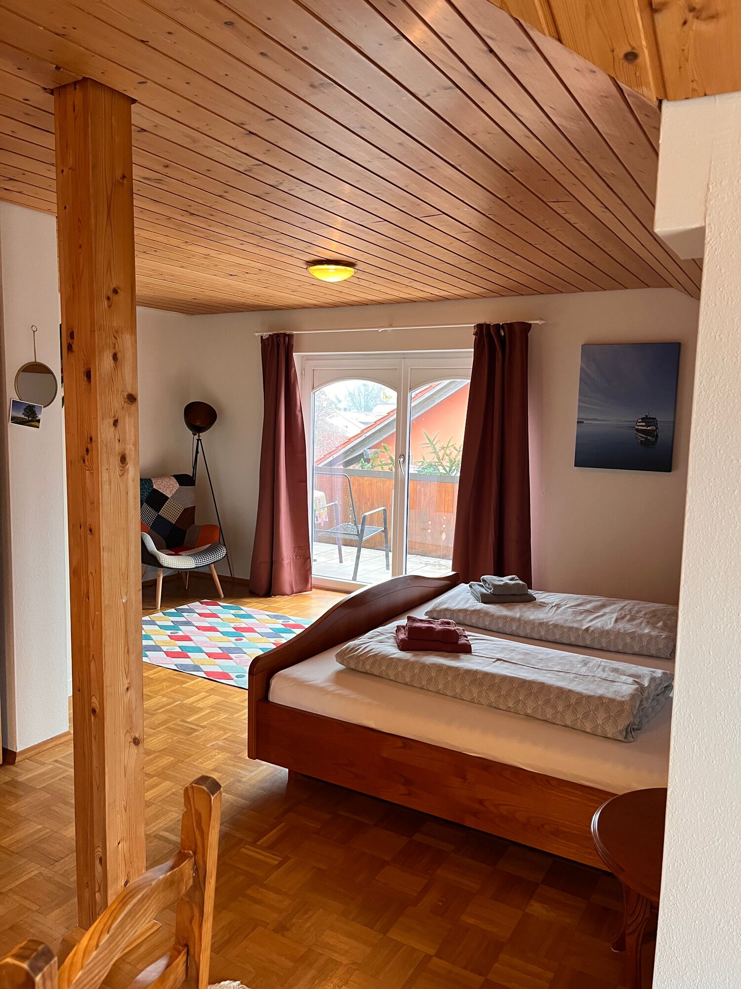 A room with a wooden ceiling, a bed with two mattresses, a chair, a floor lamp, and a view through a glass door.