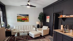 A modern living room with a sofa, armchair, ottoman, and coffee table. There is a painting on the wall, a plant, and a kitchen area with a sink and countertop.