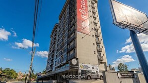 A multi-story building with a prominent red vertical sign, multiple balconies, and a clear blue sky. - Apt in Downtown Blumenau Building Une1105 (Blumenau)
