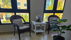 A room with two wicker chairs, a small white table, a potted plant, and large windows with a view of buildings and palm trees. - Paradise Penthouse at Aquatika Loiza (Vieques)