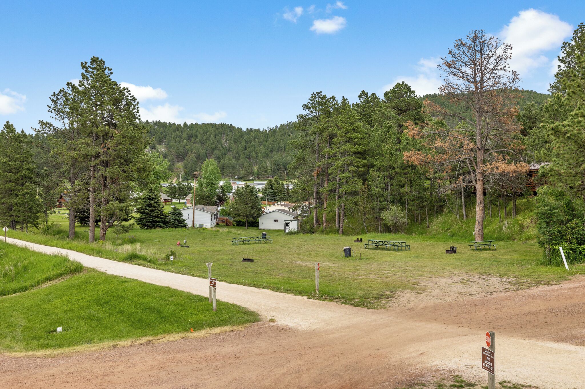 A rural setting with a dirt path, picnic tables, and cabins surrounded by trees.