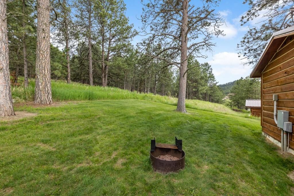 A wooden cabin with a grassy yard, a metal fire pit, and tall trees in the background.