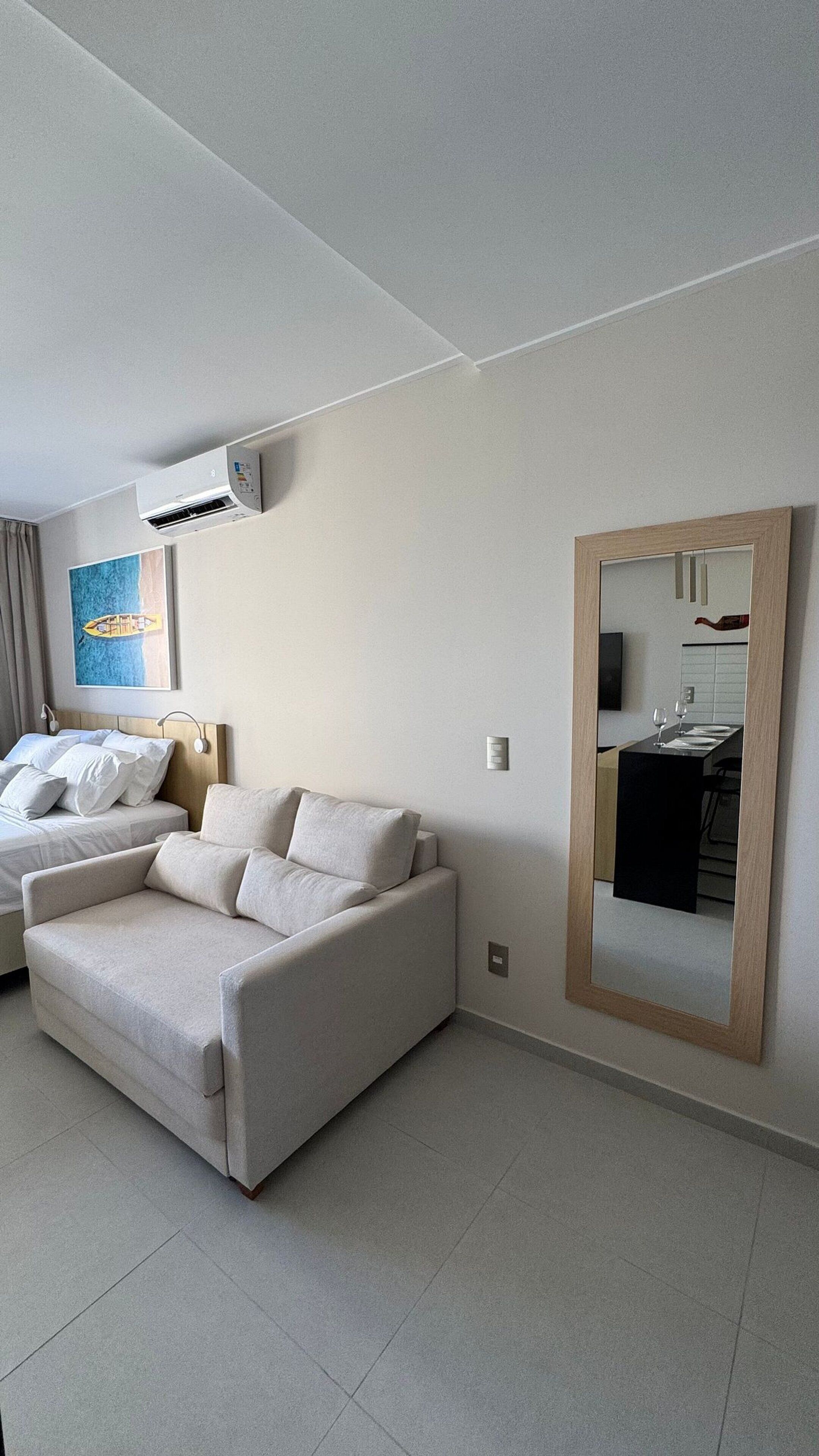 A modern hotel room with a sofa, a bed, an air conditioning unit, a painting, and a mirror reflecting a dining area.