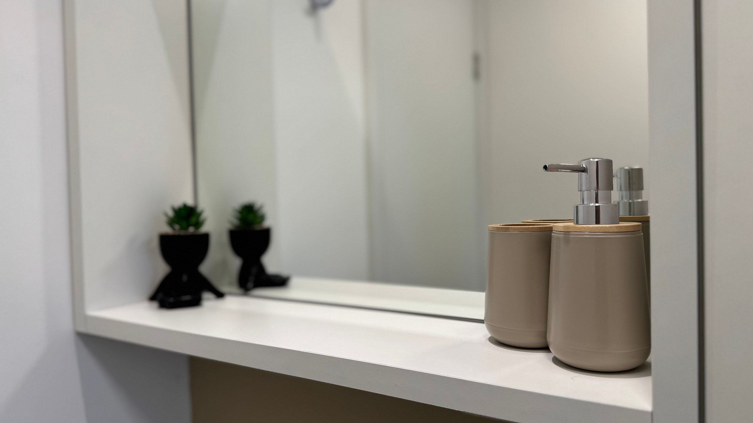 A bathroom with a mirror, two potted plants, and two beige dispensers on a white countertop.