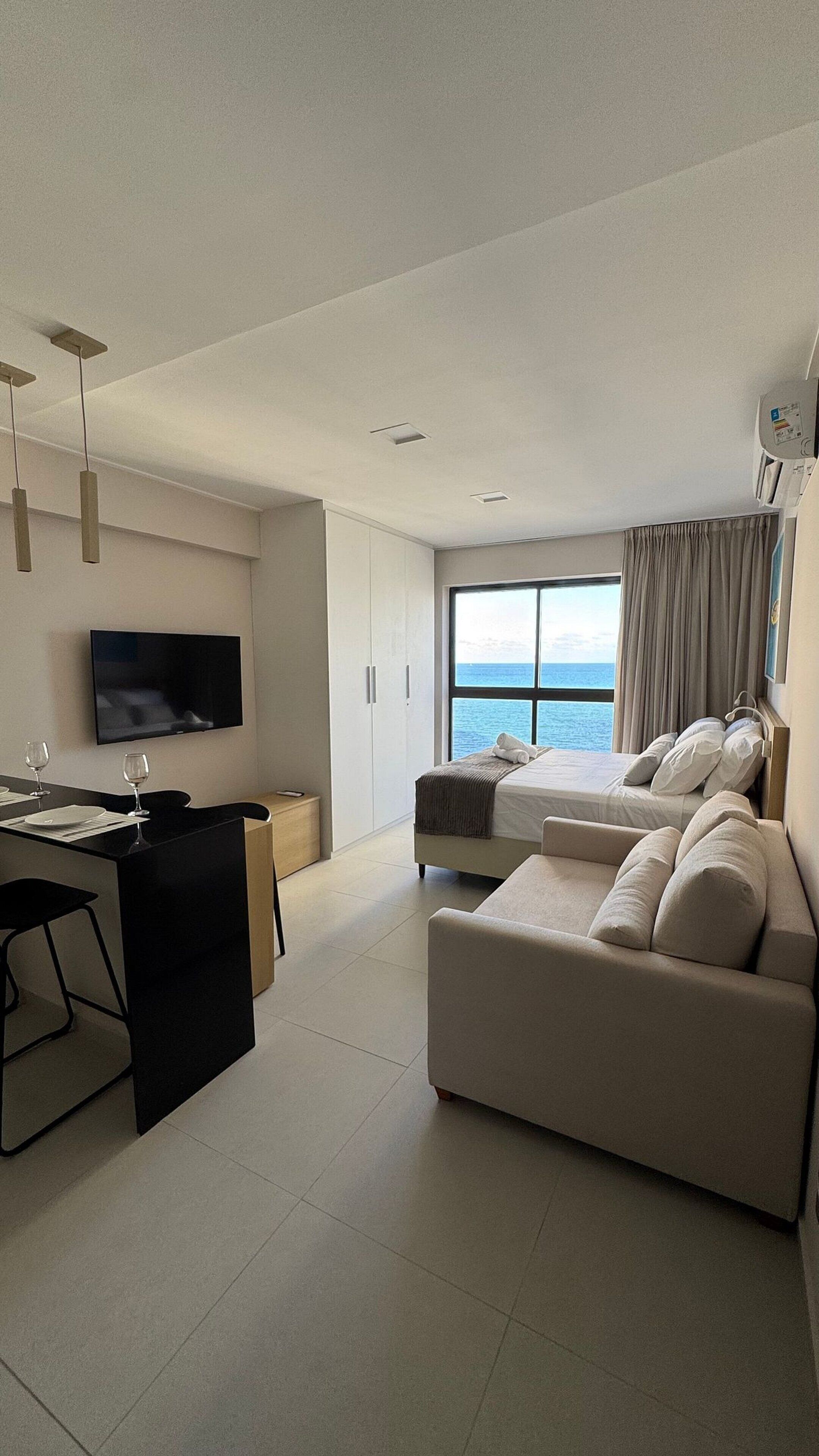 A modern hotel room with a sofa, a dining area, a TV, and a large window with a view of the sea.