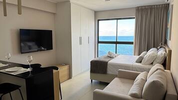 A modern hotel room with a sofa, a dining area, a TV, and a large window with a view of the sea.