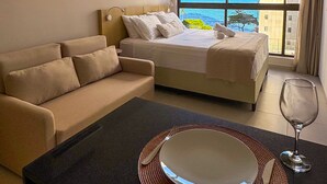 A hotel room with a bed, a sofa, a dining table with a plate and wine glass, and a large window with a view of the sea.