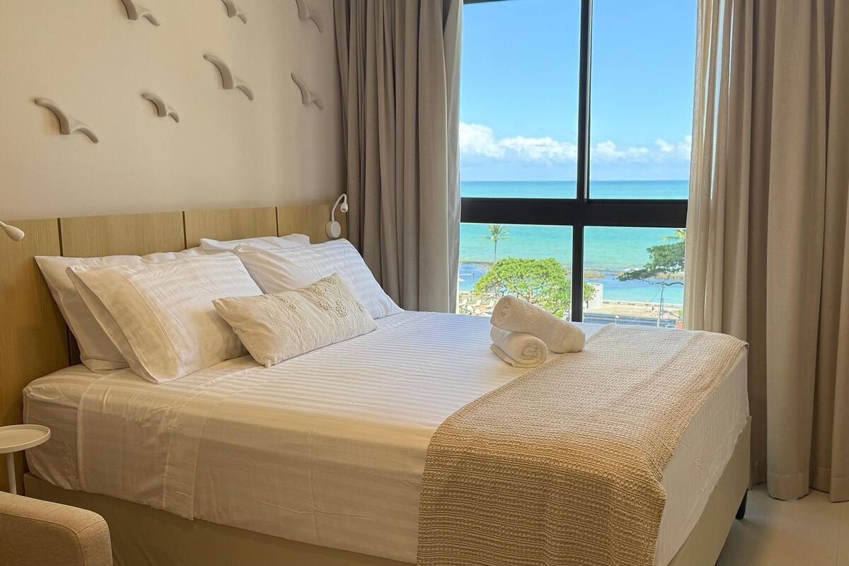 Habitación de hotel con una cama grande, vista a la playa y una ventana con cortinas.