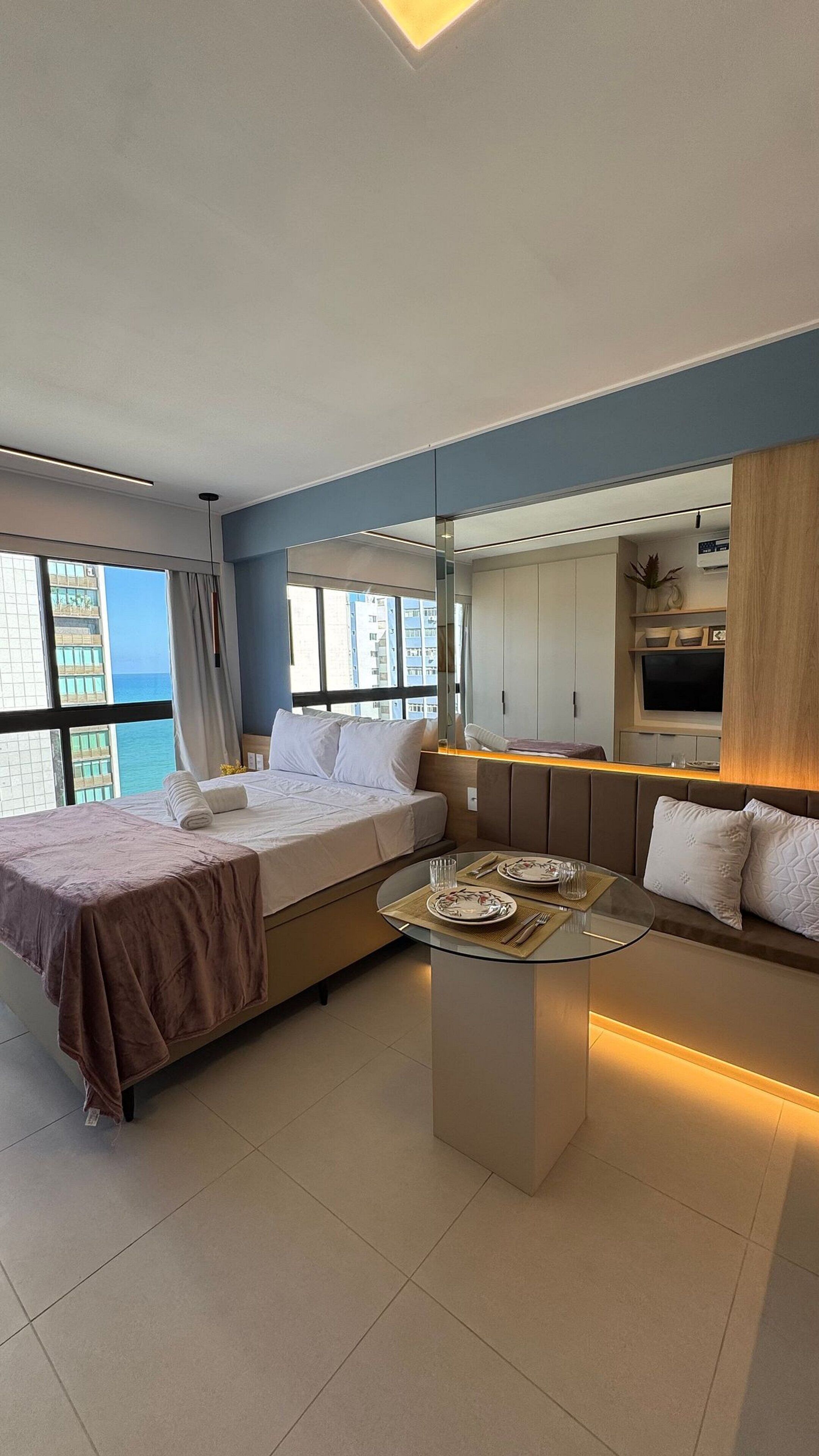 A modern hotel room with a bed, a sofa, a dining table, and a large window offering a view of the ocean.