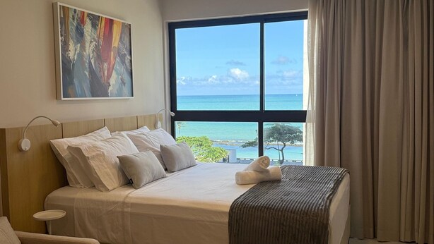 A hotel room with a bed, a painting, a window with a view of the ocean, and a curtain.